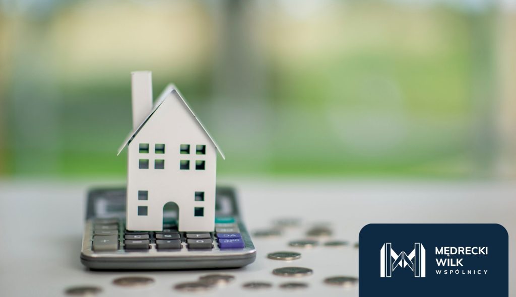 Papierowy model domu ustawiony na kalkulatorze, obok rozrzucone monety – symbol finansów i kredytów hipotecznych. W prawym dolnym rogu logo kancelarii Mędrecki Wilk i Wspólnicy