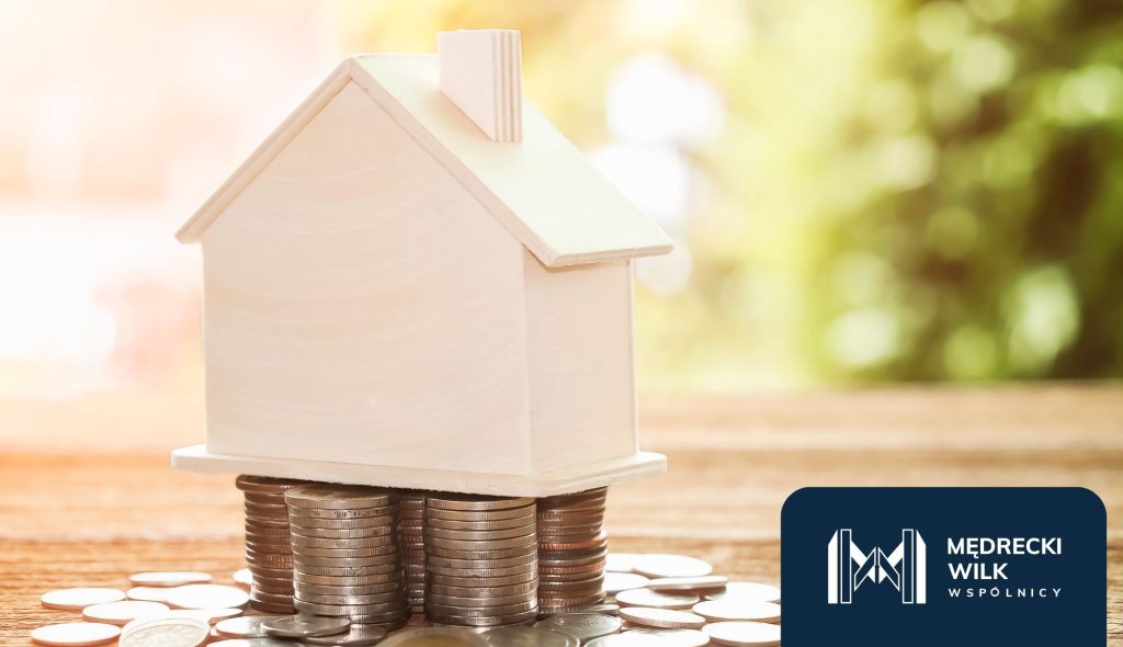 Makieta domu stojąca na stosie monet – symbol kredytu hipotecznego i finansów. W prawym dolnym rogu logo kancelarii Mędrecki Wilk i Wspólnicy.