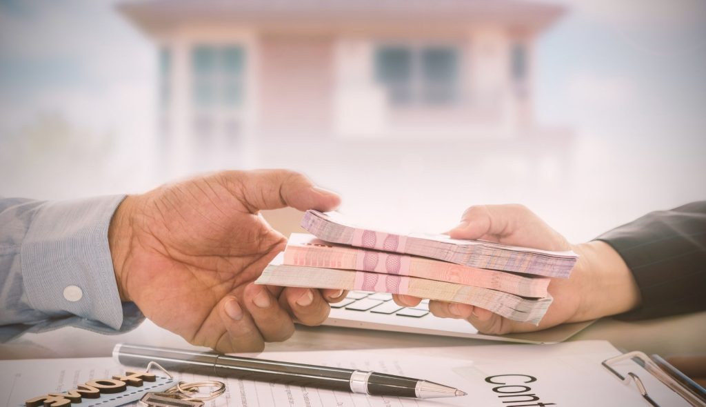 Symboliczna scena – przekazywanie pliku banknotów pomiędzy dwiema osobami nad dokumentami umowy, na stole leżą klucze i długopis