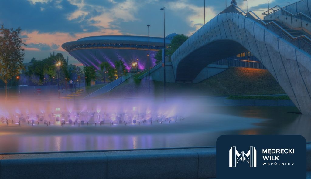katowice-kancelaria-chf • Kancelaria Mędrecki Wilk i Wspólnicy Zdjęcie przedstawia wieczorny widok Katowic — Spodek oraz most w tle, z kolorowo oświetloną fontanną na pierwszym planie. Całość utrzymana jest w chłodnych tonach niebiesko-fioletowych. W prawym dolnym rogu znajduje się logo Mędrecki Wilk i Wspólnicy.