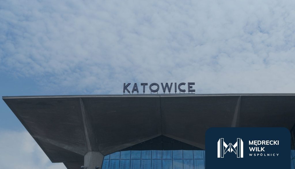 Zdjęcie przedstawia gmach dworca kolejowego w Katowicach z napisem „KATOWICE” na charakterystycznym betonowym zadaszeniu. Niebo jest częściowo zachmurzone, a w prawym dolnym rogu znajduje się logo Mędrecki Wilk i Wspólnicy.