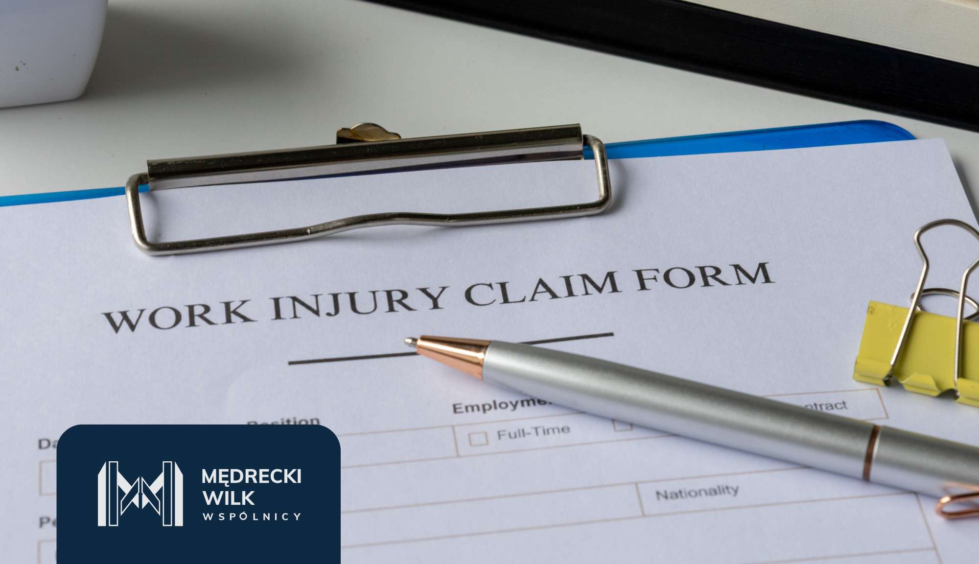 Close-up of a Work Injury Claim Form on a desk with a silver pen and metal binder clip nearby, and a blue clip edge visible; company logo overlay in the corner.