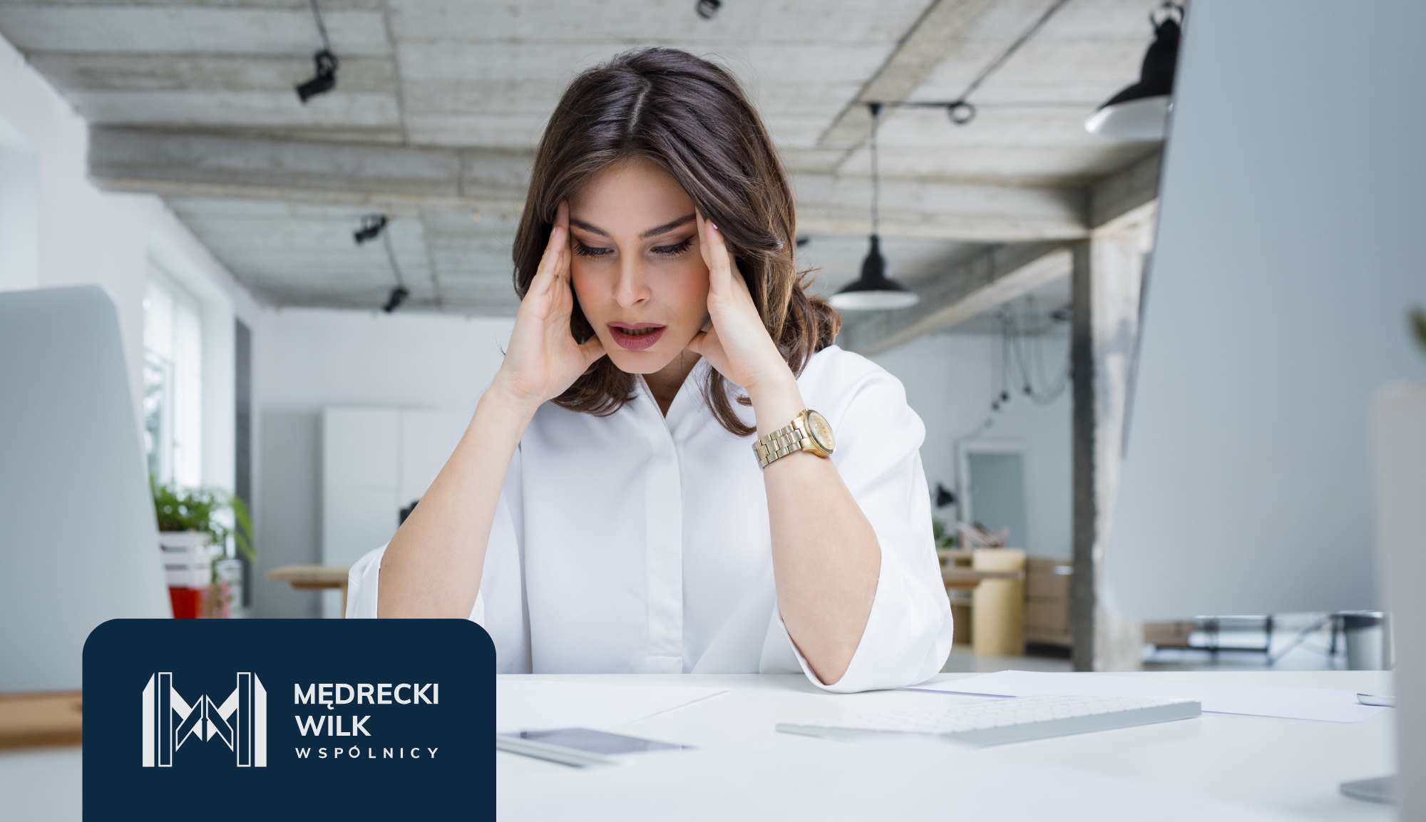 Professional woman in a white blouse sits at a desk in a modern office, hands on temples and a stressed expression.