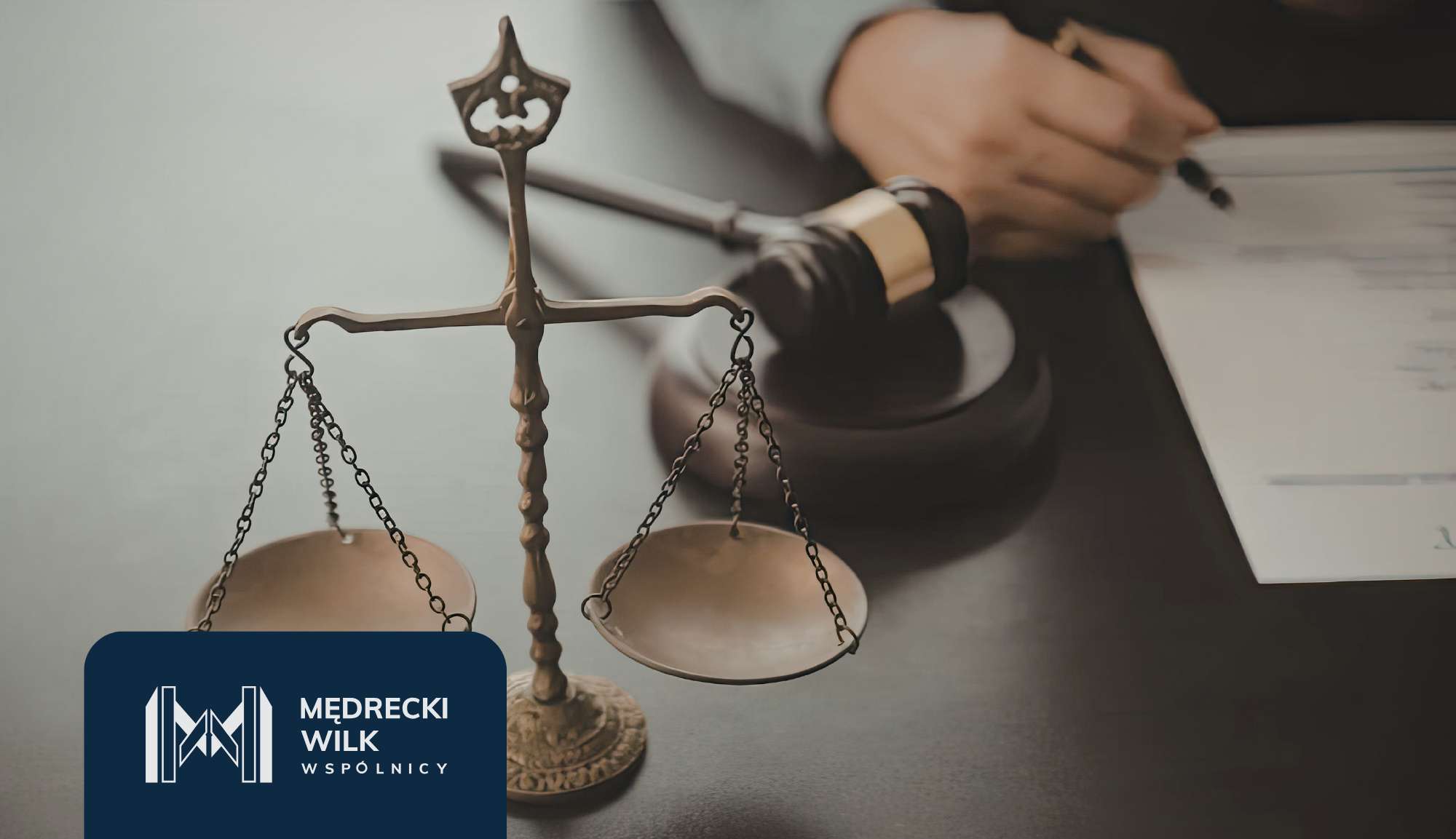 Bronze scales of justice in focus with a blurred person signing documents in the background; law firm branding visible at the bottom left.