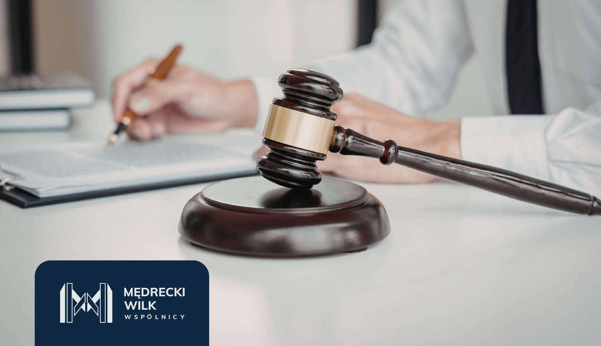 Close-up of a judge's gavel on a desk with a blurred person signing documents in the background; law firm logo in the bottom-left corner.