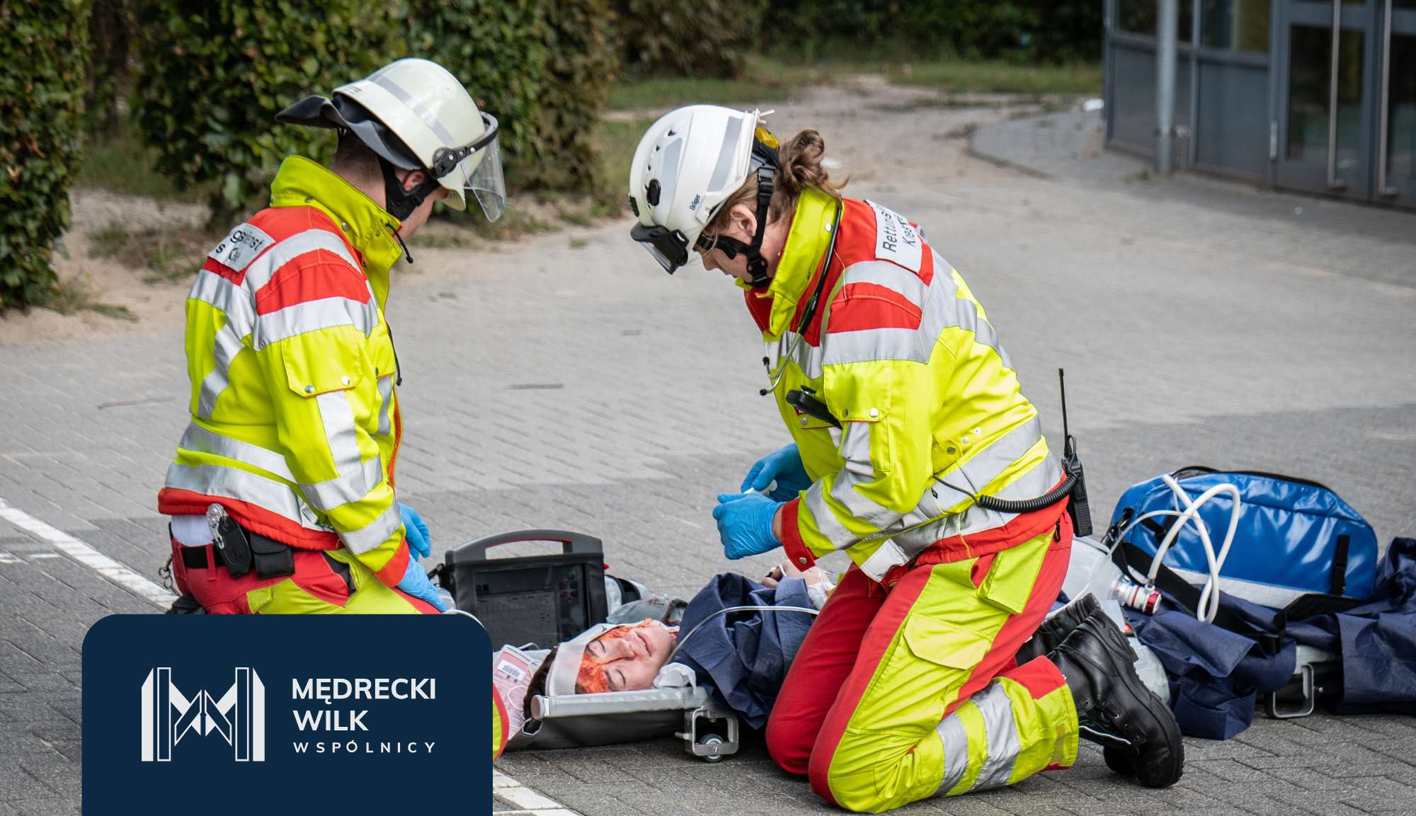 Two emergency responders in high-visibility gear attend to an injured person on a paved surface, with medical equipment nearby.