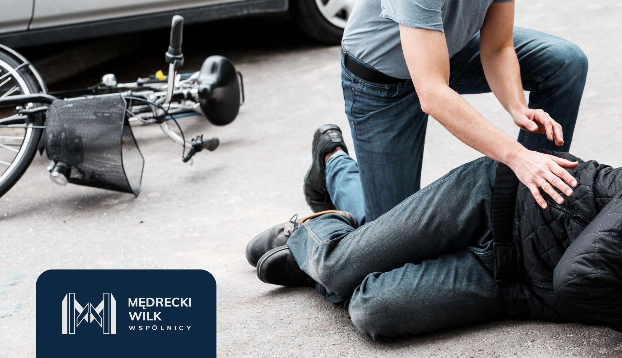 Person kneeling beside a fallen cyclist, assessing an injured rider on a paved surface; bicycle lies nearby with a basket visible.