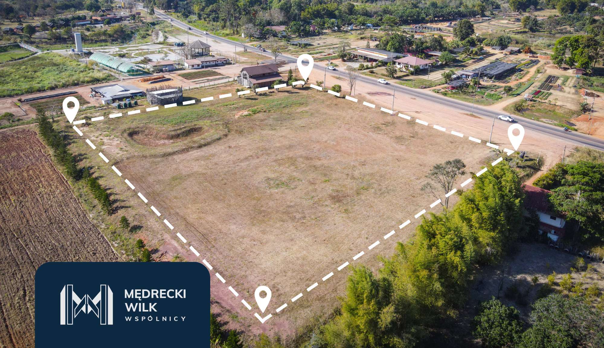 Aerial view of a large vacant plot outlined by white dashed lines with three map-pin markers, near a road and small buildings in a rural town. Logo overlay in bottom-left corner.