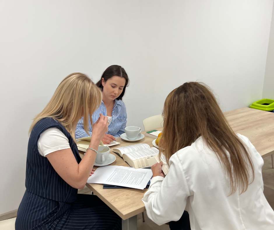 Three women at a wooden table studying with open books and coffee cups.