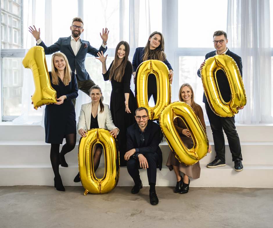 Group of colleagues celebrating a milestone with gold '1000' balloons in a bright office setting.