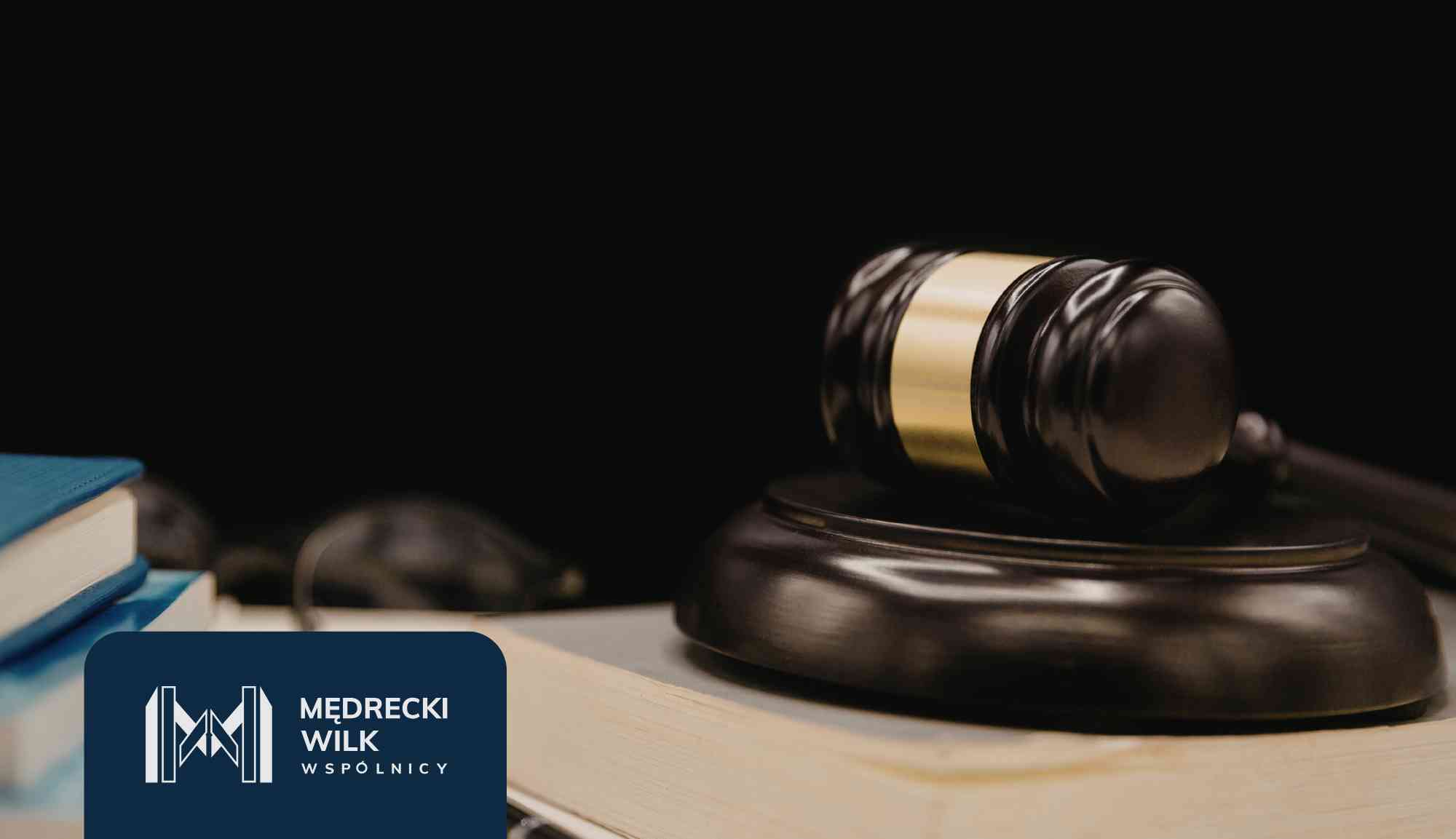 Bronze scales of justice in focus with a blurred person signing documents in the background; law firm branding visible at the bottom left.