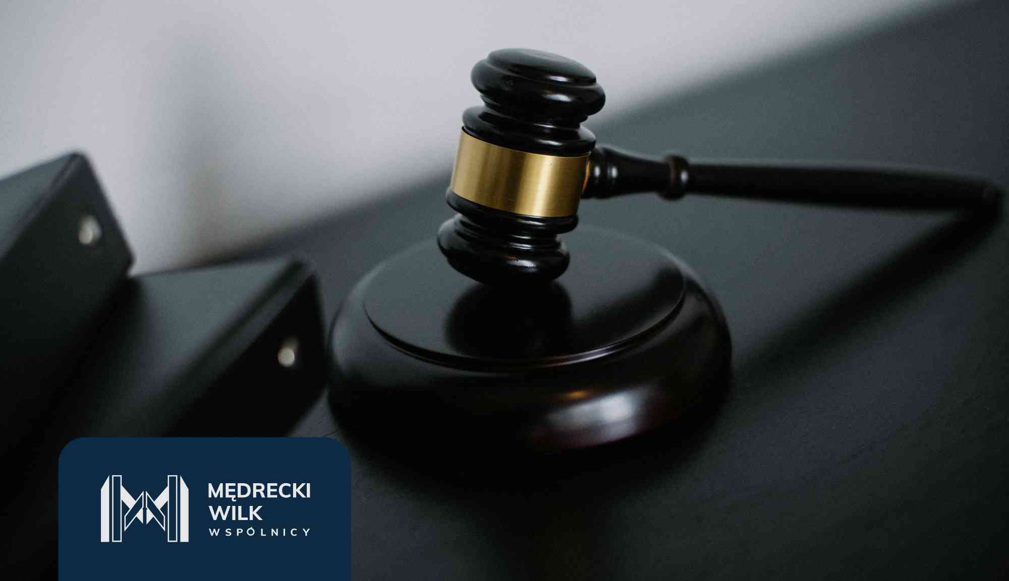 Close-up of a wooden gavel with a gold band resting on a dark desk, with a blue-law firm logo in the corner.