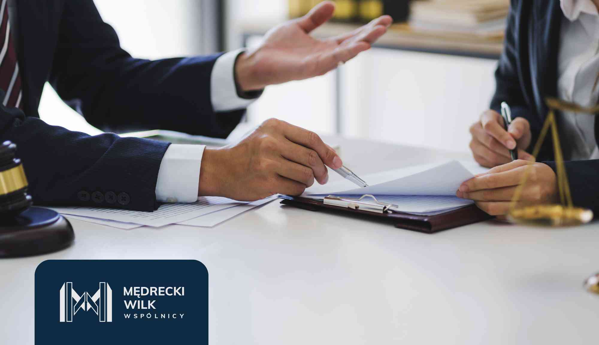 Two professionals in business attire sign documents at a desk, with a blue logo panel visible in the foreground.