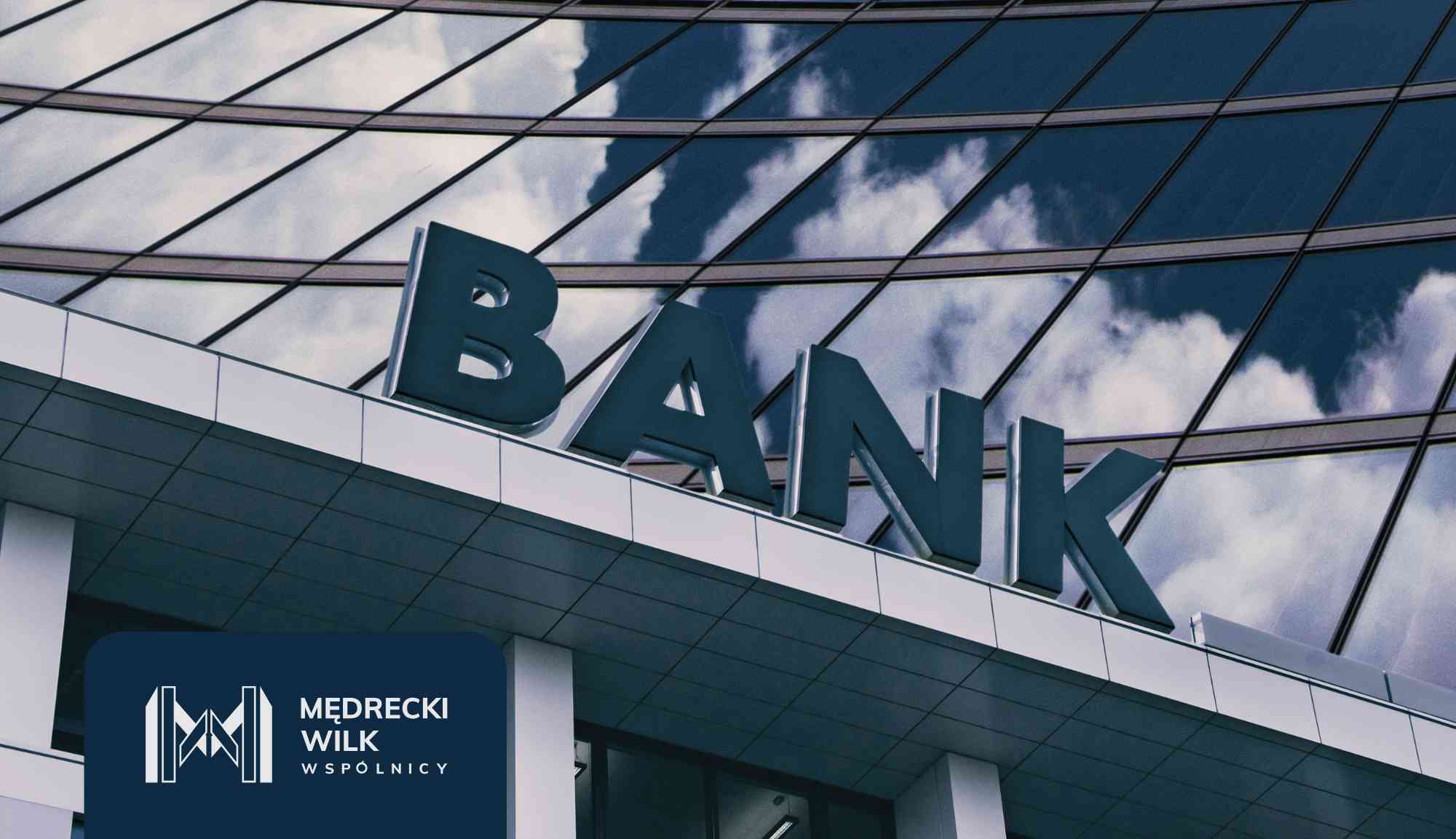 Modern glass office building with the word 'BANK' on the facade and a dark blue panel showing the Medrecki Wilk logo and 'WSPÓLNICY'.