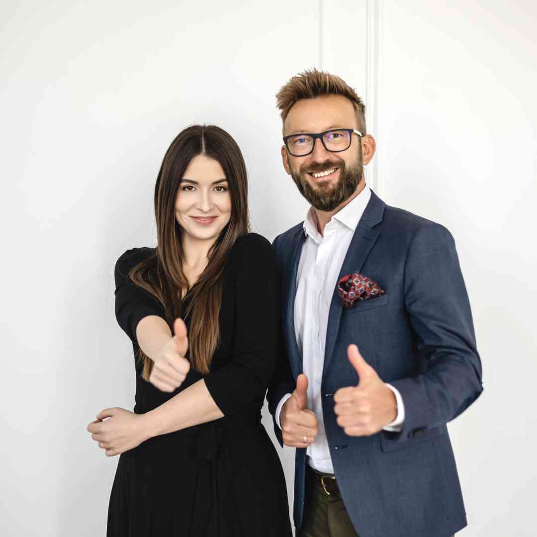 Smiling man in a blue blazer and white shirt with pocket square and a woman in black dress giving thumbs up. (two people)