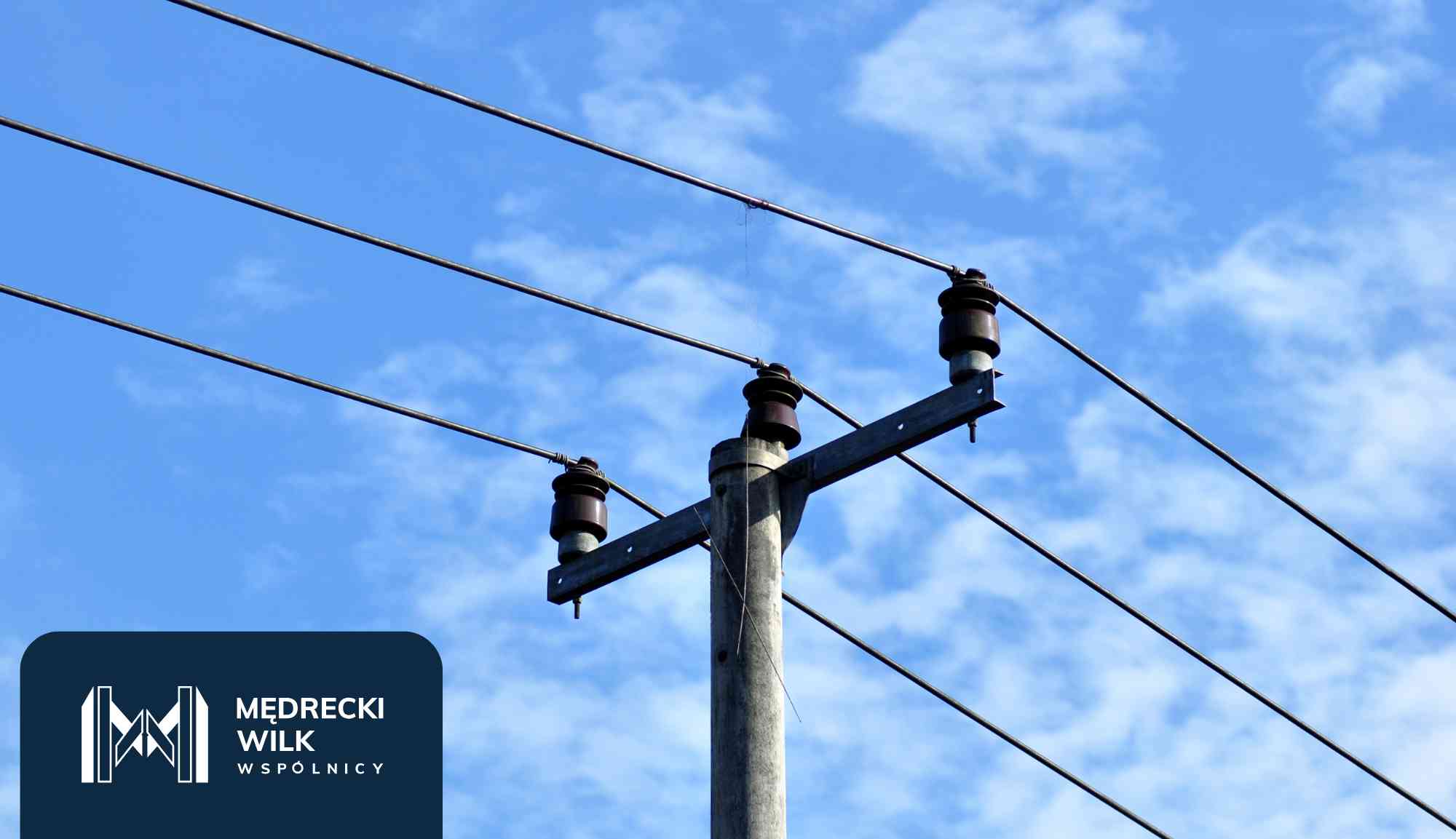 Power lines and tall transmission towers silhouetted against a colorful sunset sky, with a dark blue overlay and company logo in the lower-left corner reading 'Medrecki Wilk Wspólnicy'
