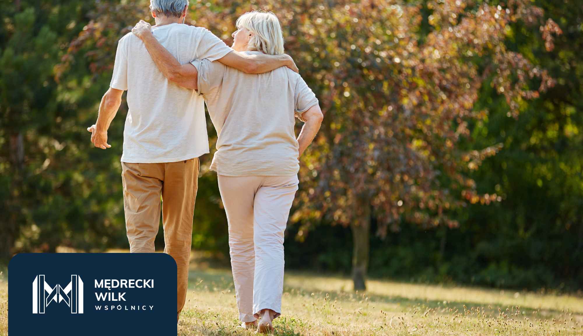 Older couple walking arm-in-arm through a sunlit park, with branding in the bottom-left corner.
