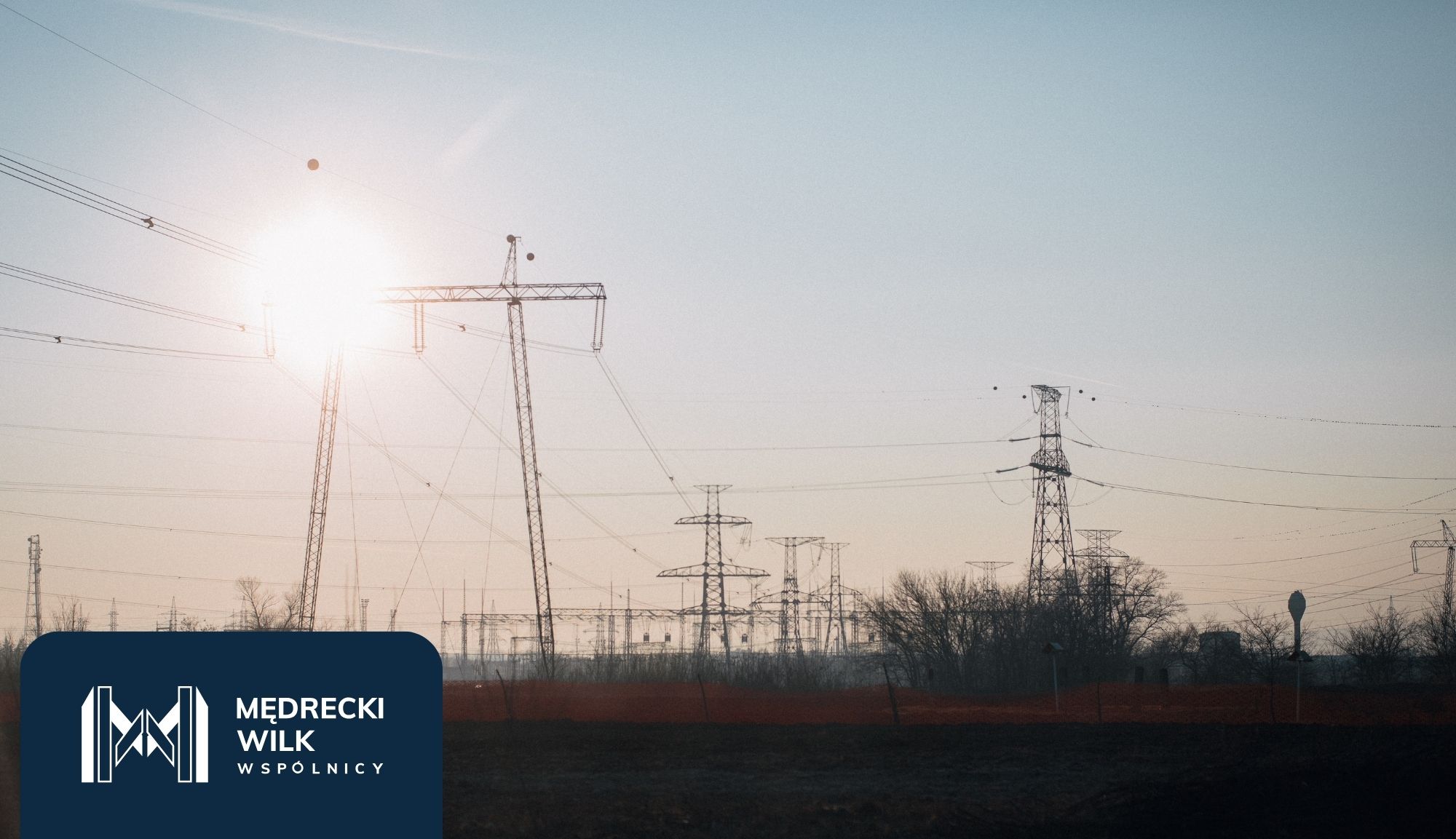 Power lines and tall transmission towers silhouetted against a colorful sunset sky, with a dark blue overlay and company logo in the lower-left corner reading 'Medrecki Wilk Wspólnicy'