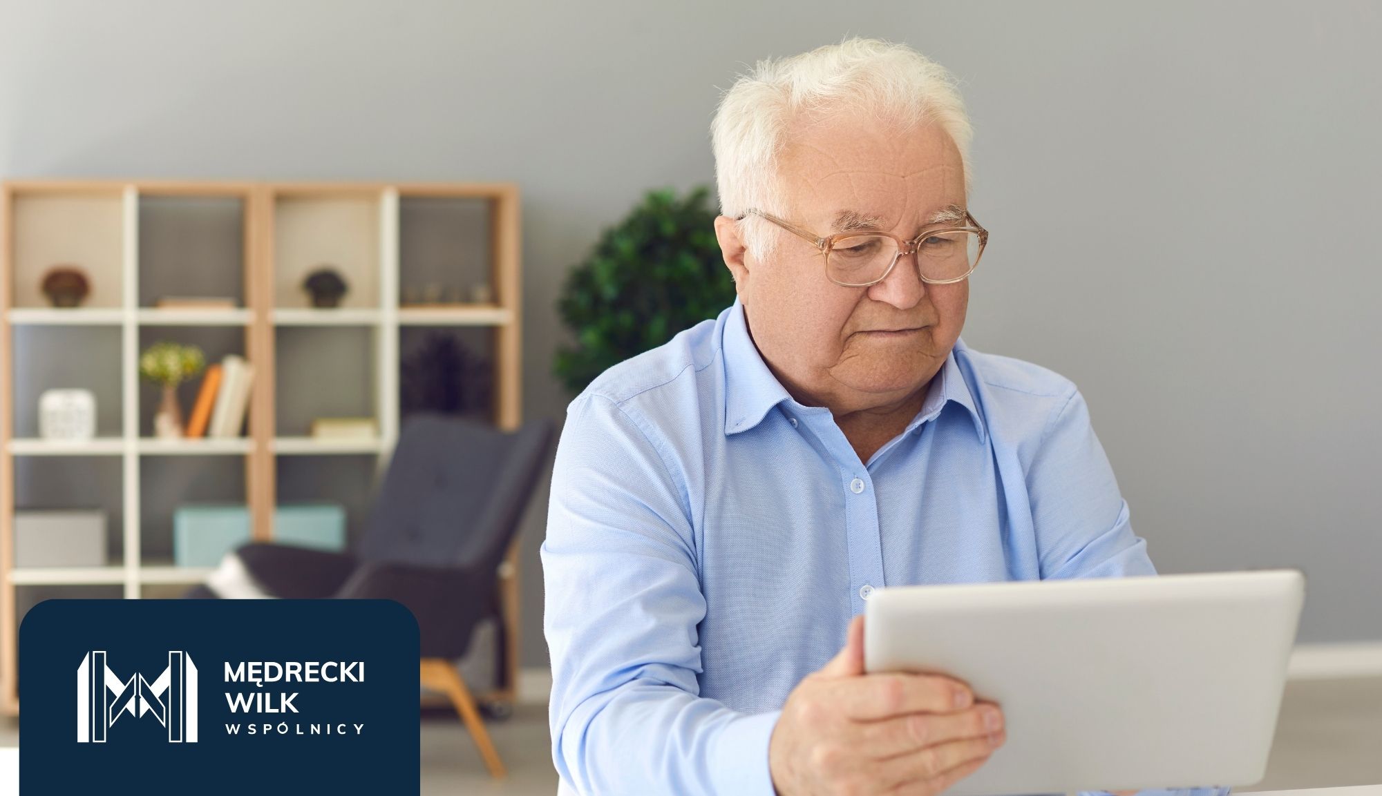 Older man with white hair and glasses in a blue shirt using a tablet at a desk in a modern office, branding panel visible bottom-left.