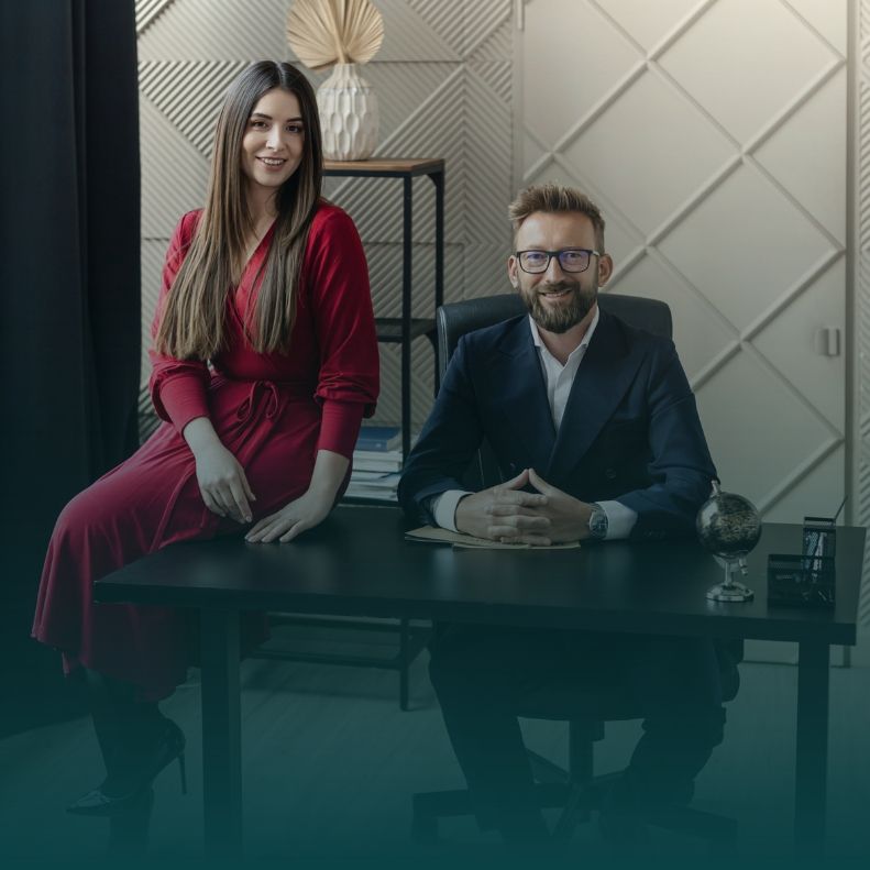 medrecki-wilk-kontakt Two professionals sit at a dark desk in a modern office, smiling at the camera: a woman in red and a man in a suit with glasses.